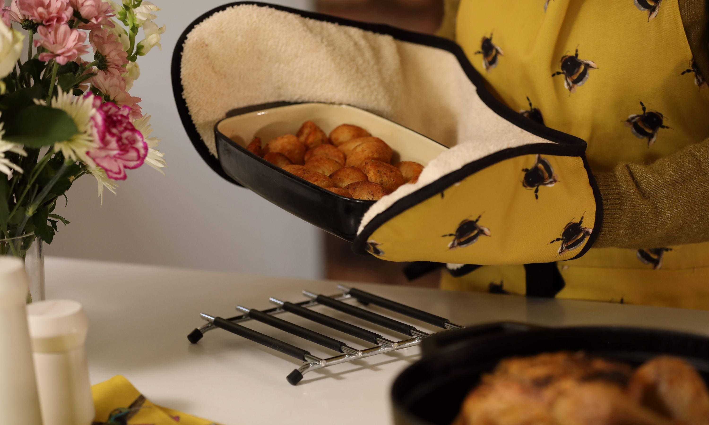 Person holding some roast potatoes using a bee oven glove whilst wearing a bee apron in a lovely bright yellow