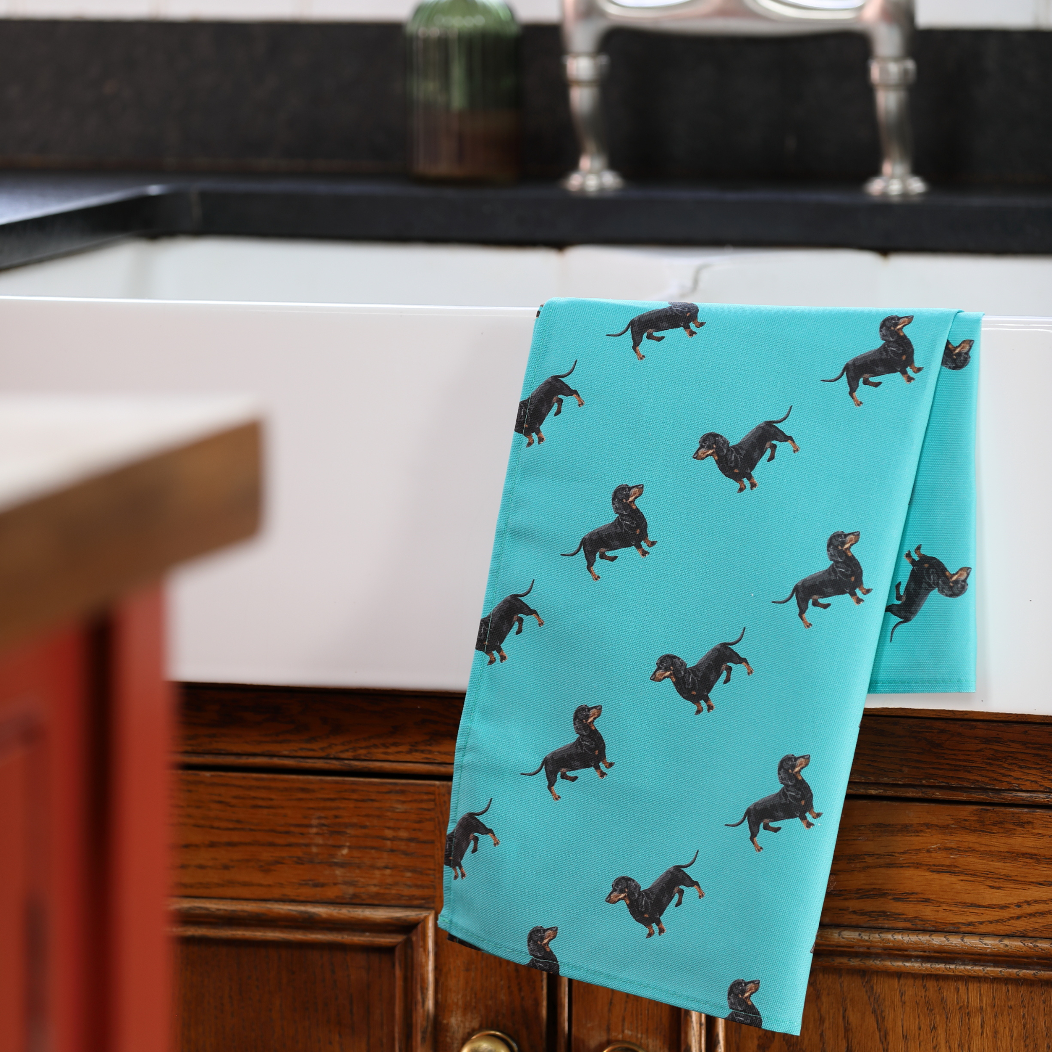 A turquoise kitchen towel with a repeating pattern of black and tan dachshunds draped over the edge of a white farmhouse sink in a kitchen setting.