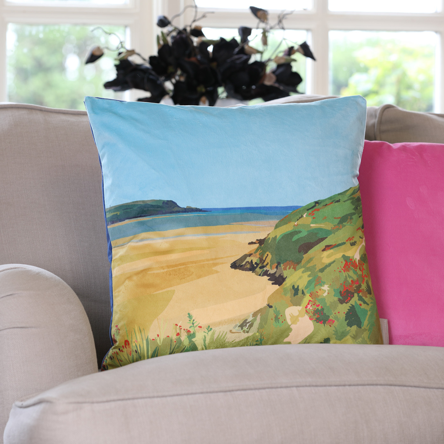 A velvet cushion with a printed design featuring a beach and hillscape, placed on a sofa with a floral cushion behind it.
