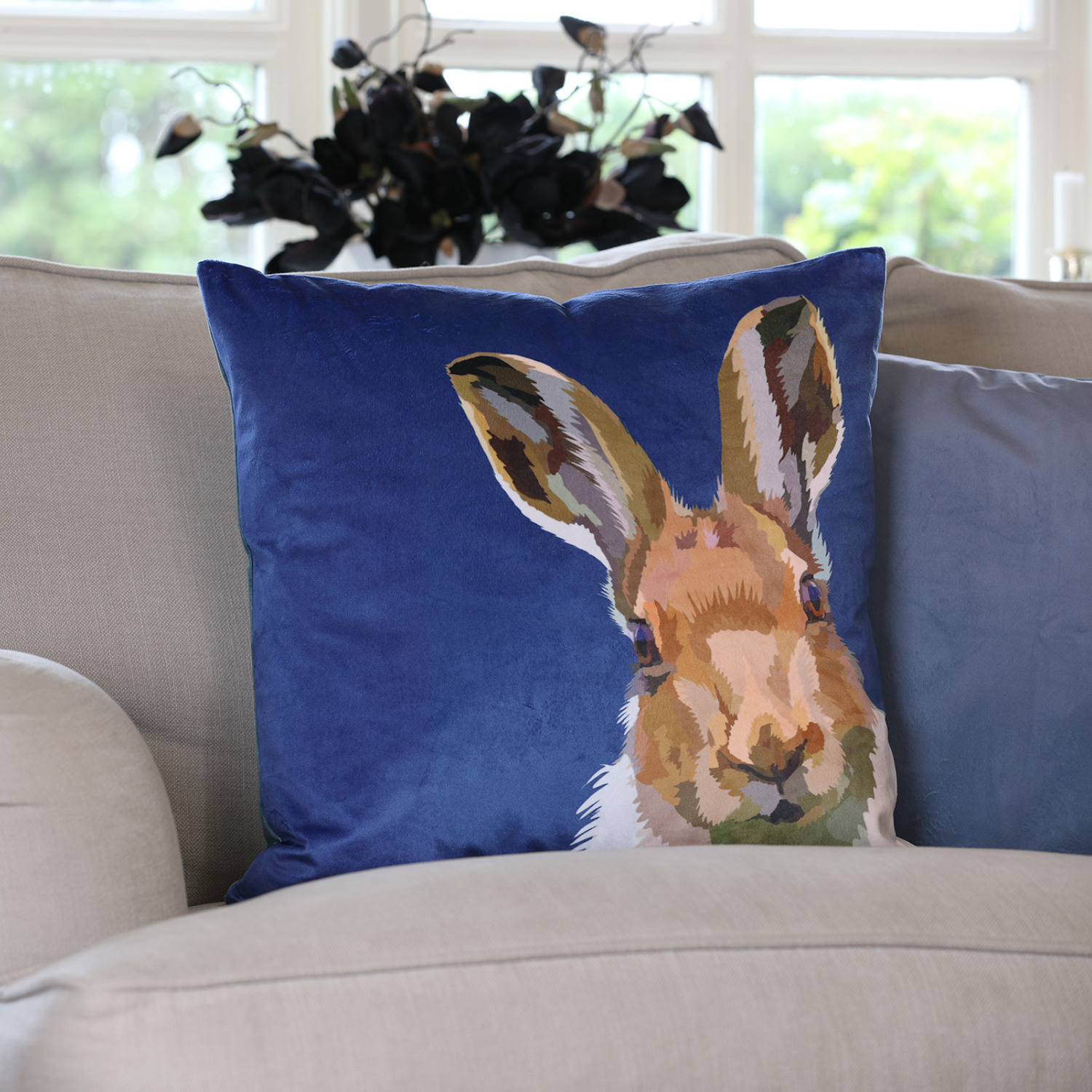 A blue velvet cushion featuring an illustration of a hare with black tipped ears on a beige sofa in a living room setting.