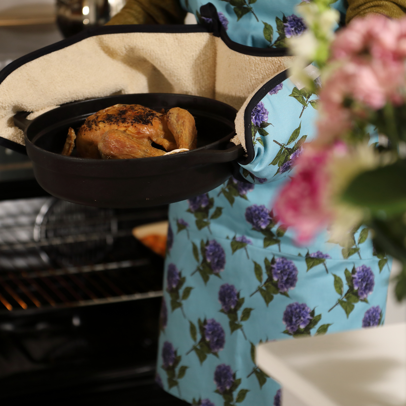 Person wearing a floral apron with a roasted chicken in a pan on a grill.