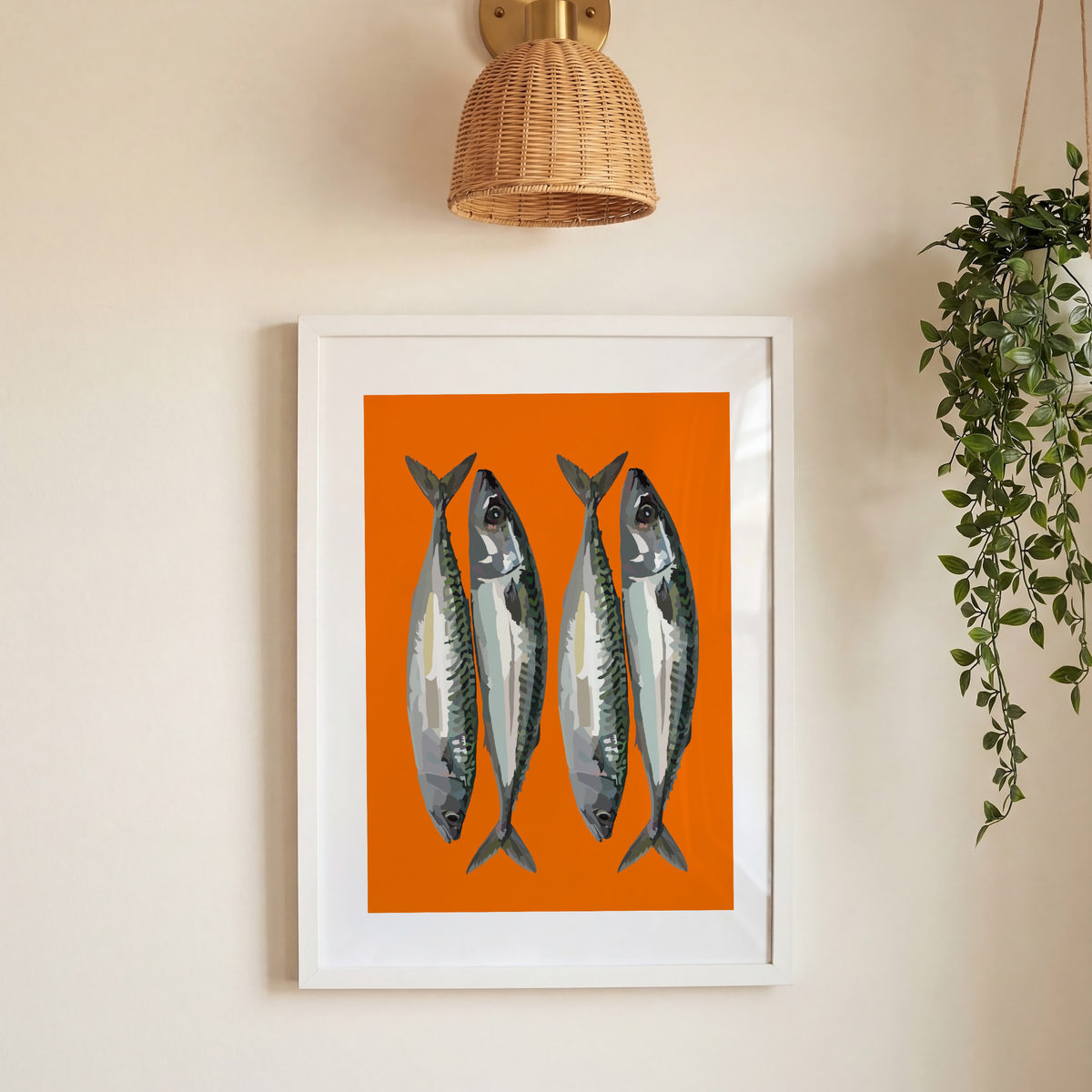 Framed wall art featuring four silver mackerel fish on a bright orange background, hung on a beige wall below a woven lampshade sconce and next to a hanging green plant.