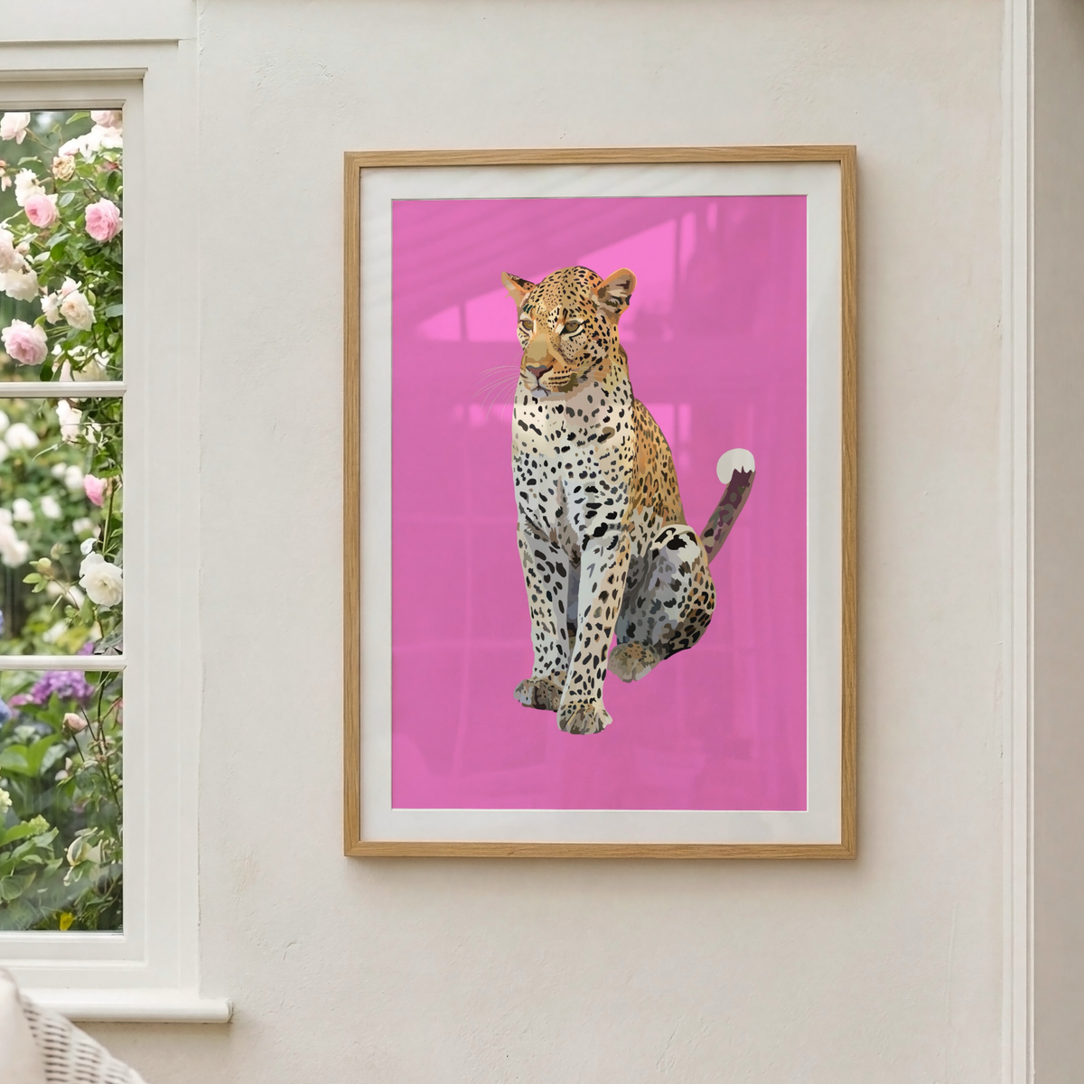 A framed art print featuring a realistic leopard cutout on a bright pink background, hanging on a light beige wall next to a window with pink and white flowers.