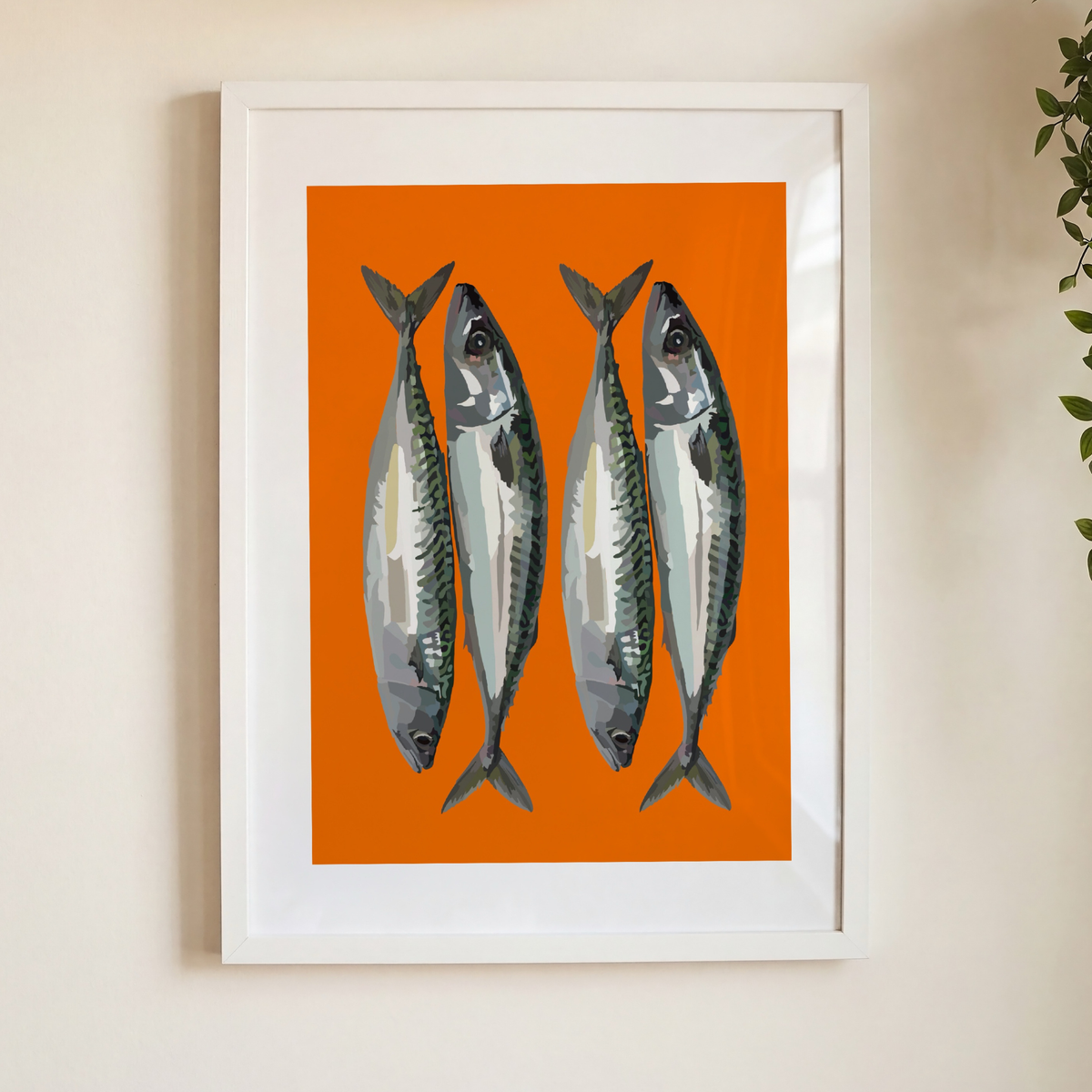 Framed wall art featuring four silver mackerel fish on a bright orange background, hung on a light beige wall with a small plant leaf visible at the right edge.