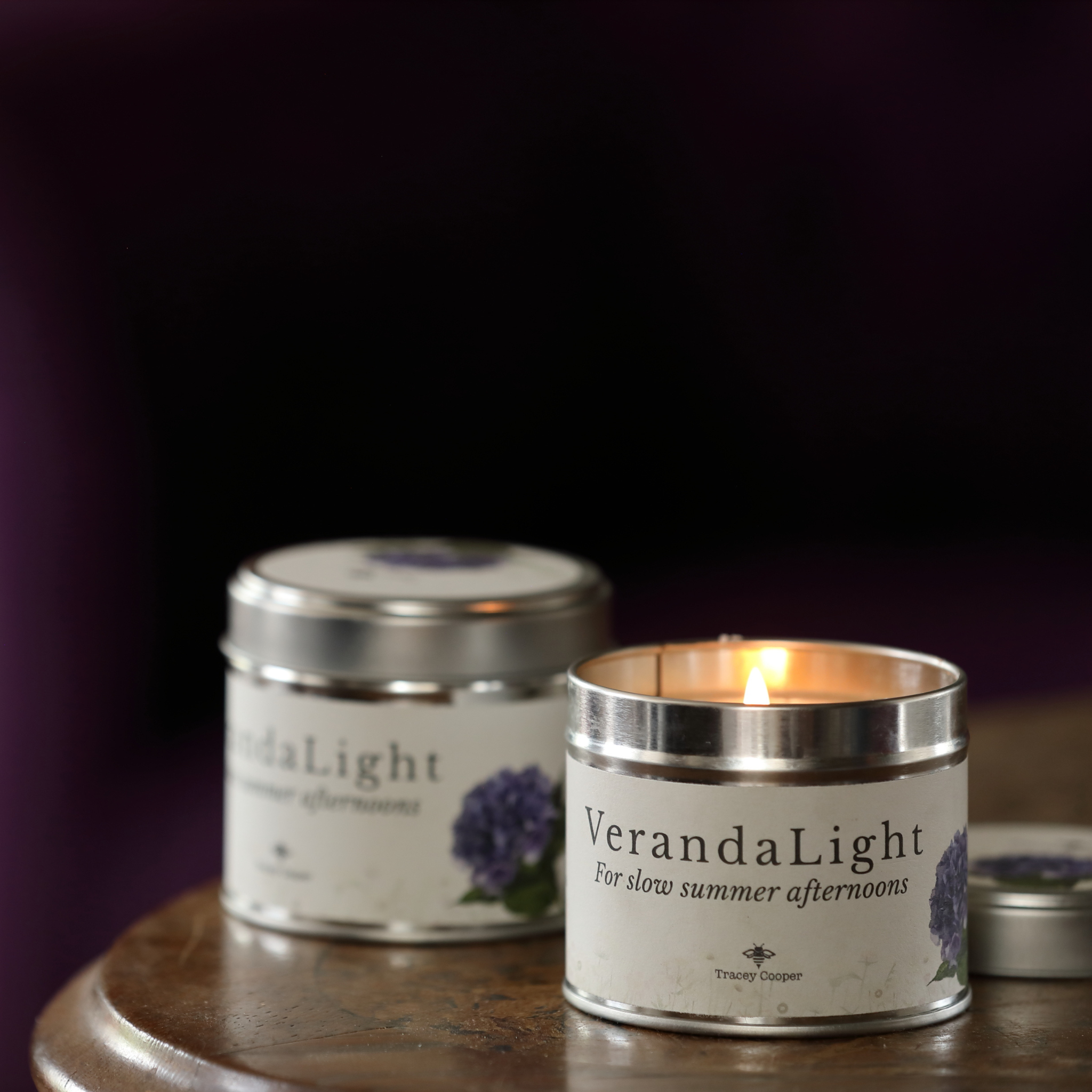 Two silver metal tin candles labeled 'VerandaLight' sit on a wooden surface against a dark purple background. One tin is lit with a warm flame, and the other is slightly out of focus behind it.