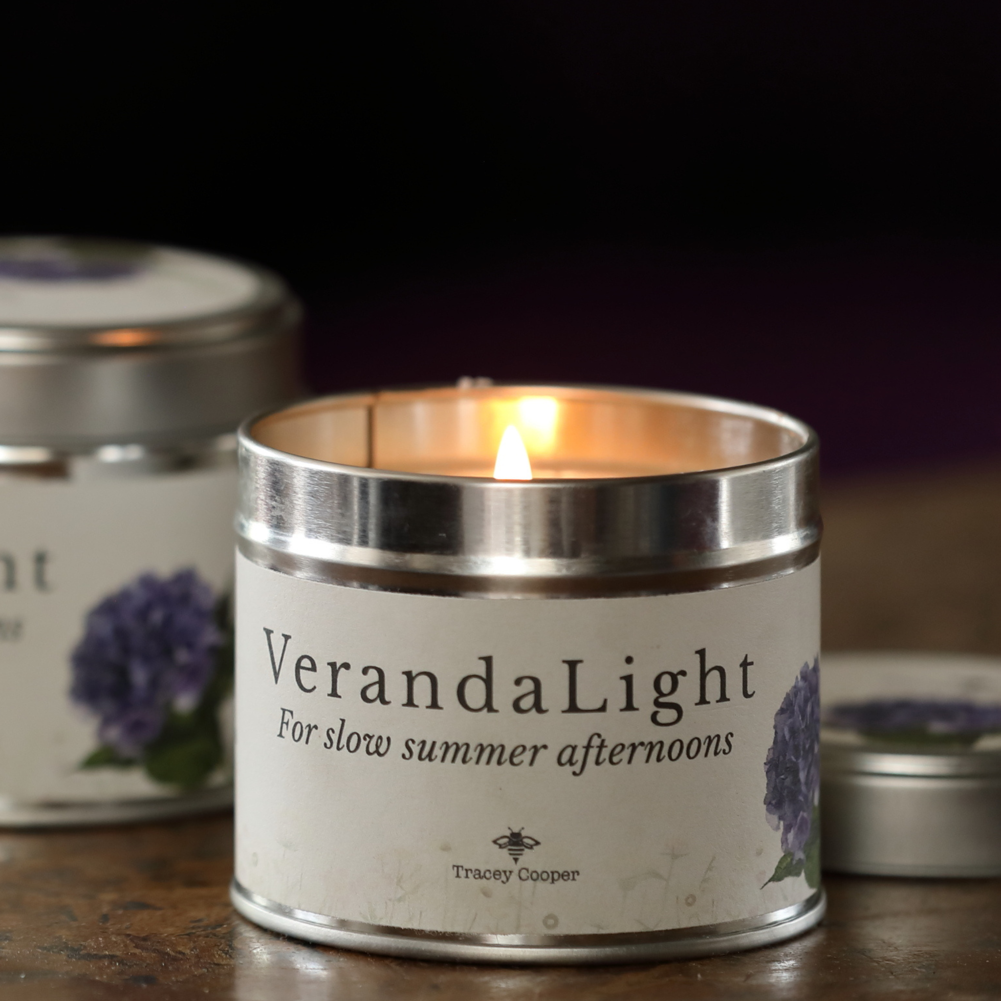 A lit silver tin candle labeled 'VerandaLight' sits on a wooden surface with a warm flame visible inside. The white label features purple floral artwork and the brand name 'Tracey Cooper' near the bottom.