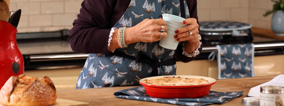 A person in a kitchen wearing a blue apron with a white tree pattern, holding a white mug. In the foreground on a wooden counter is a red baking dish with a baked dish, a loaf of bread, and other kitchen items.