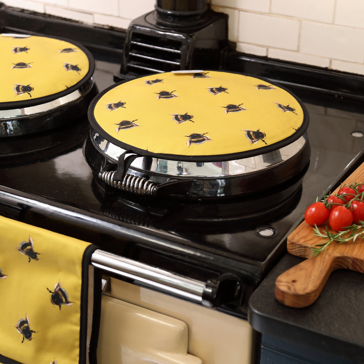 Yellow AGA pads with a black bee pattern, shown on a black stove top and hanging on a cream-colored oven handle in a kitchen setting.