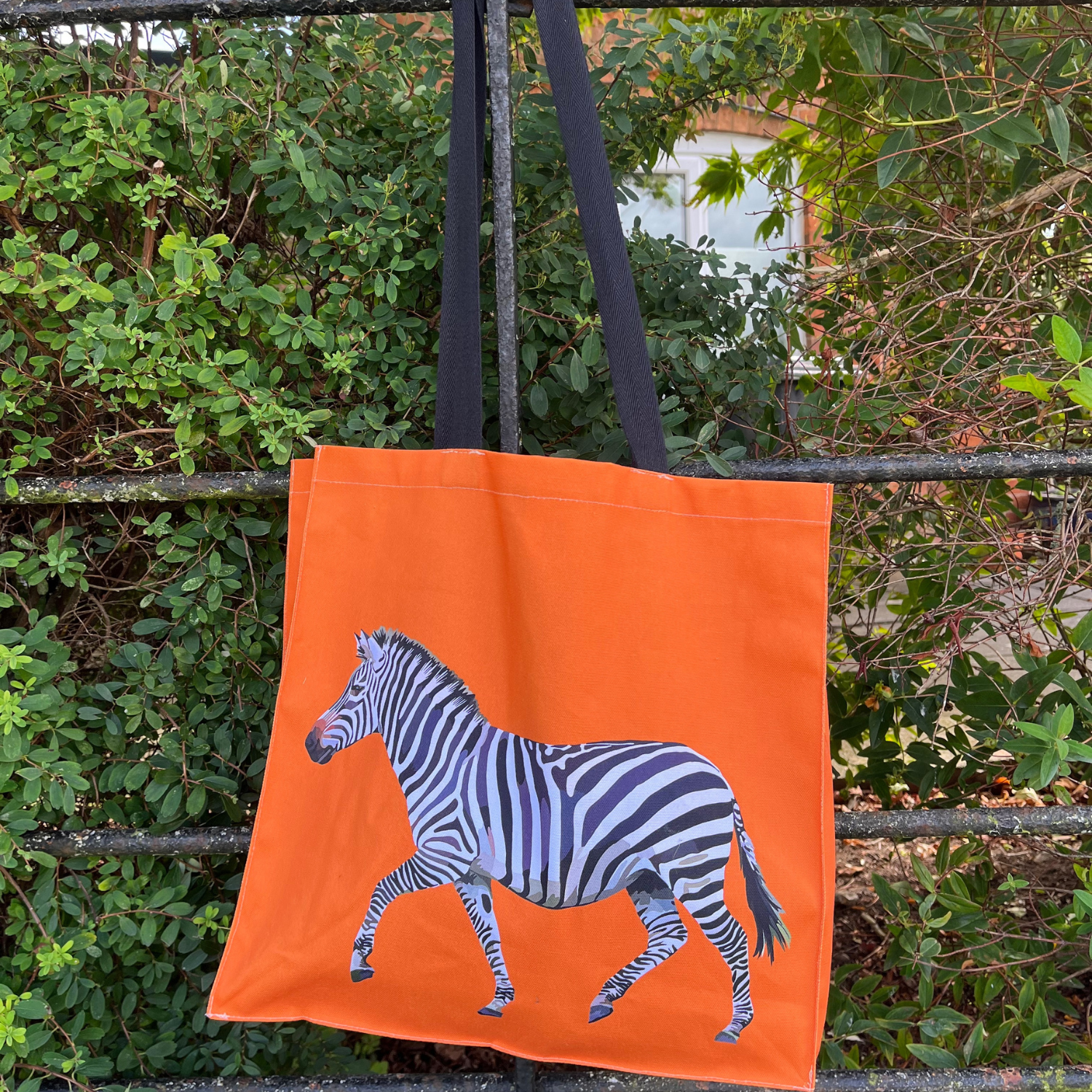 A vibrant orange tote bag featuring a black and white zebra illustration on the front, with a repeat pattern on the back, hanging on a metal rack with green foliage in the background.