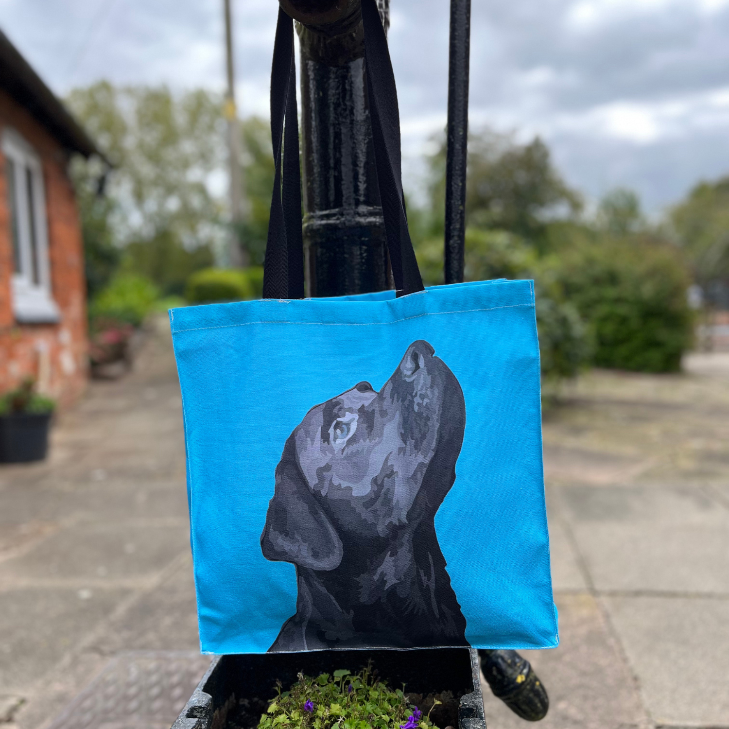 A blue tote bag with a front illustration of a black lab dog's head, hanging on a metal pole with a plant beside it.