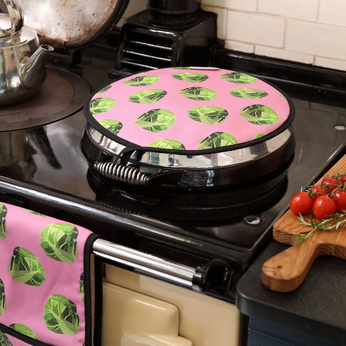 A pink aga cover with a green sprout pattern sits on a Aga stove in a kitchen setting. A matching pink ovenglove with the same sprout print hangs on the stove handle, and a wooden cutting board with red cherry tomatoes is visible on the counter.