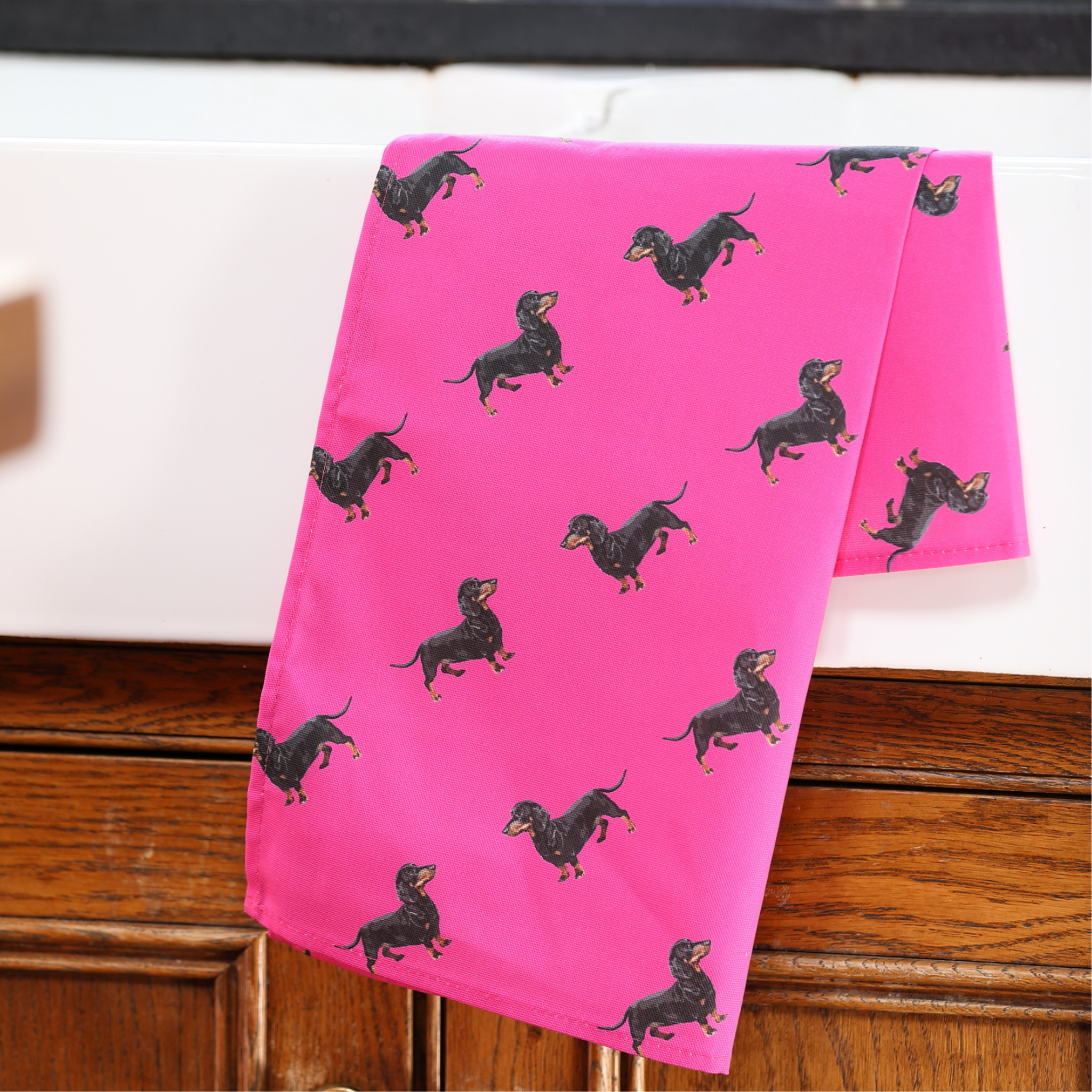 A bright pink tea towel printed with a repeating pattern of black dachshund dogs, draped over a white surface and a wooden cabinet.