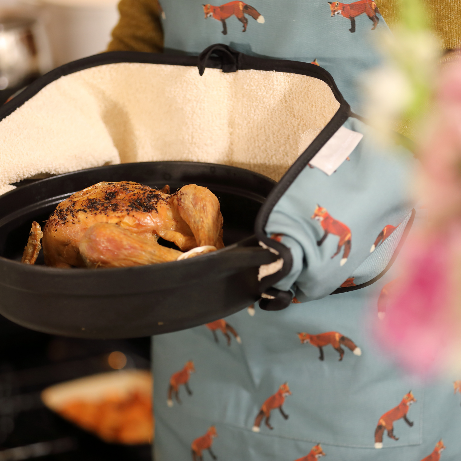 Roasted chicken in a pan held by a person wearing a blue apron with fox patterns.