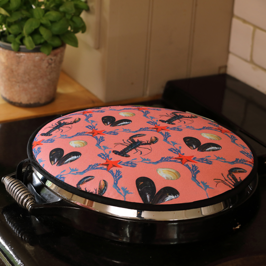 A hob cover with a lobster, mussel, and starfish design on a salmon pink background, placed on a stove.