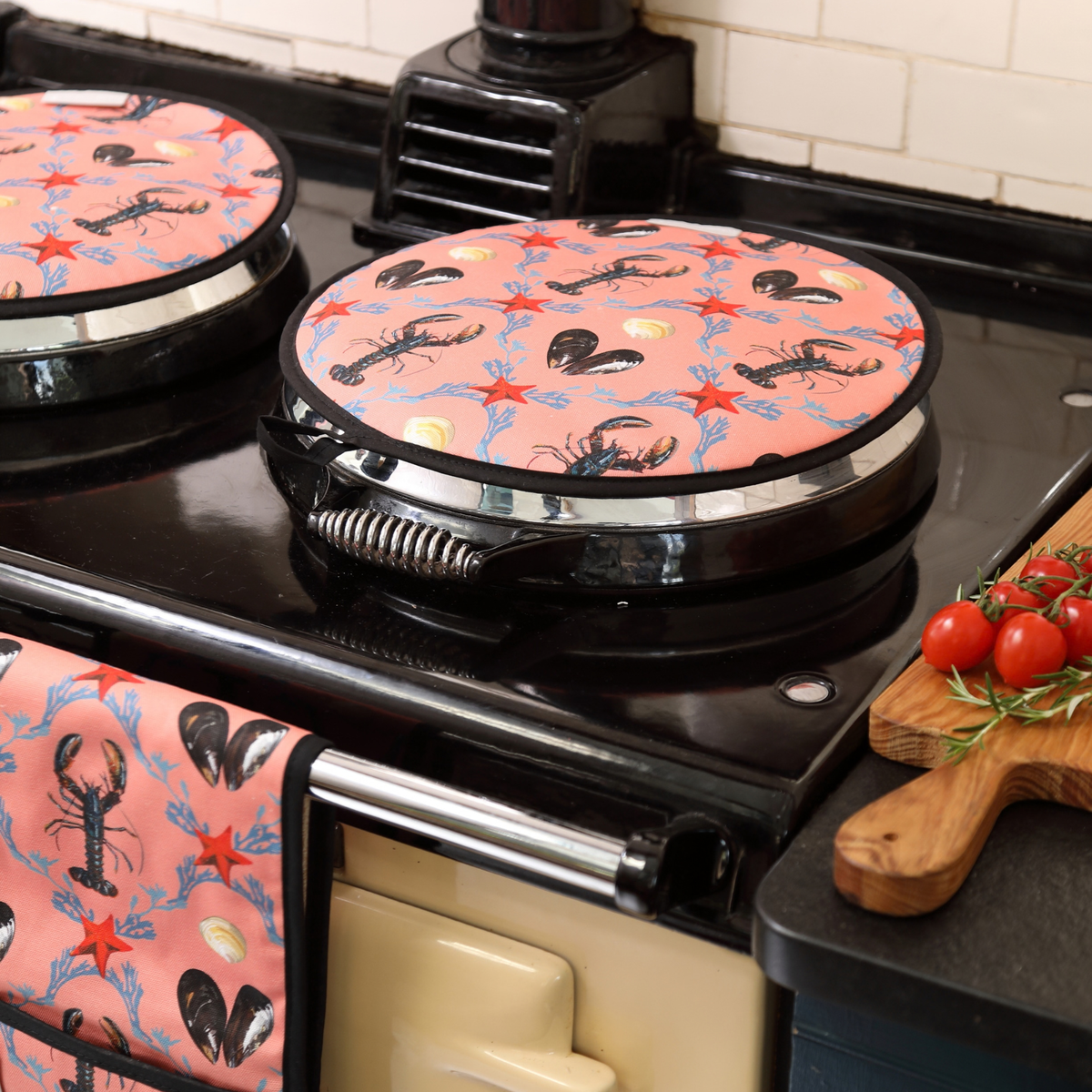 A  pink lobster-patterned AGA cover sits on a AGA  in a kitchen. A matching patterned towel hangs from the stove handle, and tomatoes are visible on a wooden cutting board to the right.