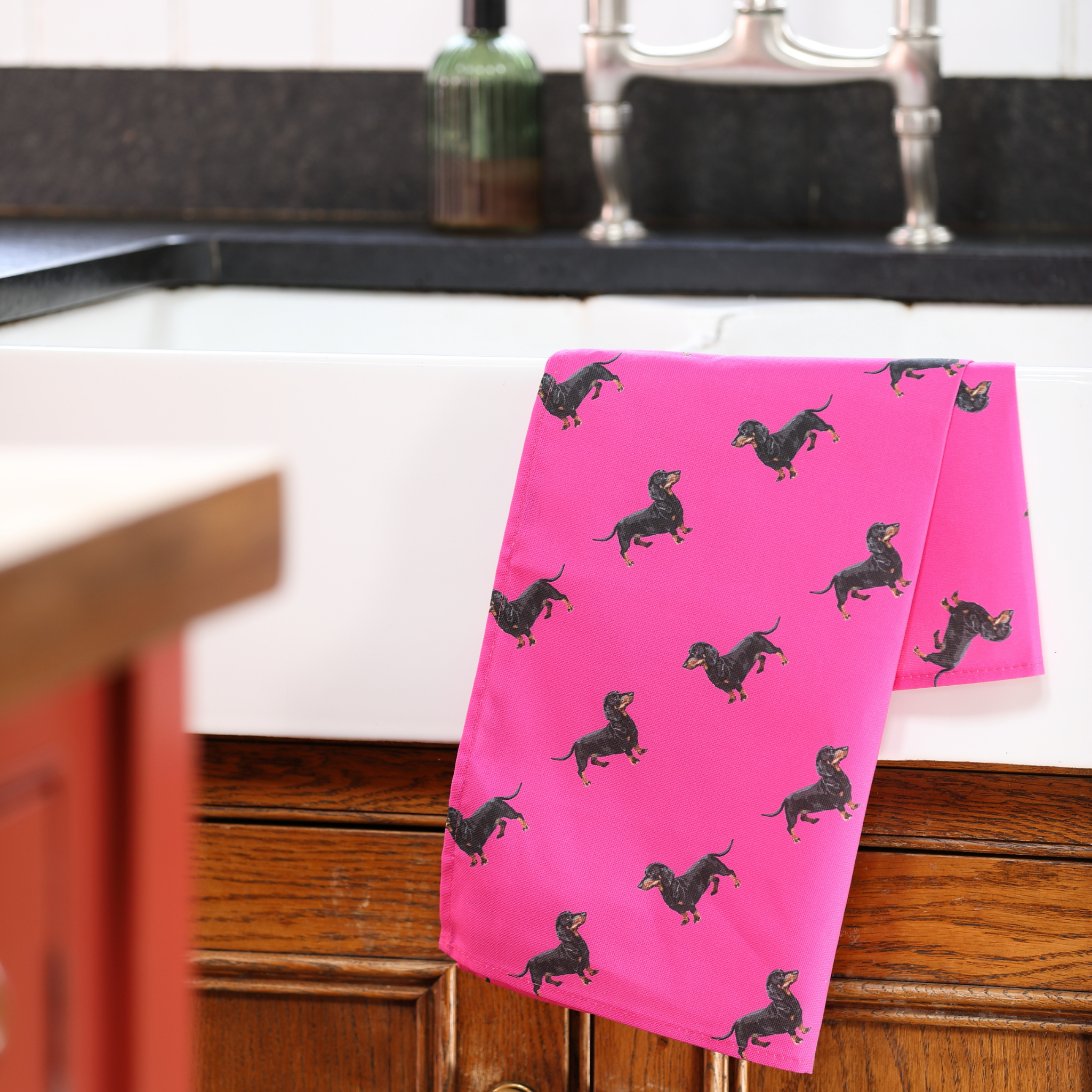 A bright pink kitchen towel with a black dachshund dog print pattern, draped over the edge of a white farmhouse sink in a kitchen setting.