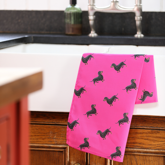 A bright pink kitchen towel with a black dachshund dog print pattern, draped over the edge of a white farmhouse sink in a kitchen setting.
