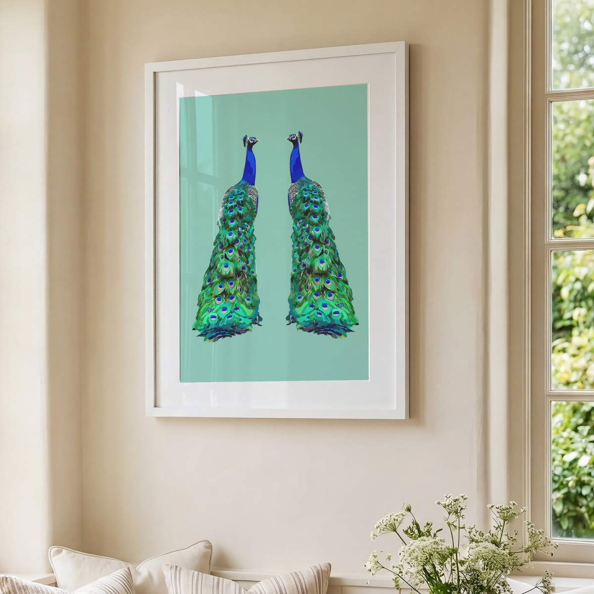 Framed wall art featuring two mirrored peacock necks and tails on a mint green background, hung on a beige wall beside a window with outdoor greenery visible.