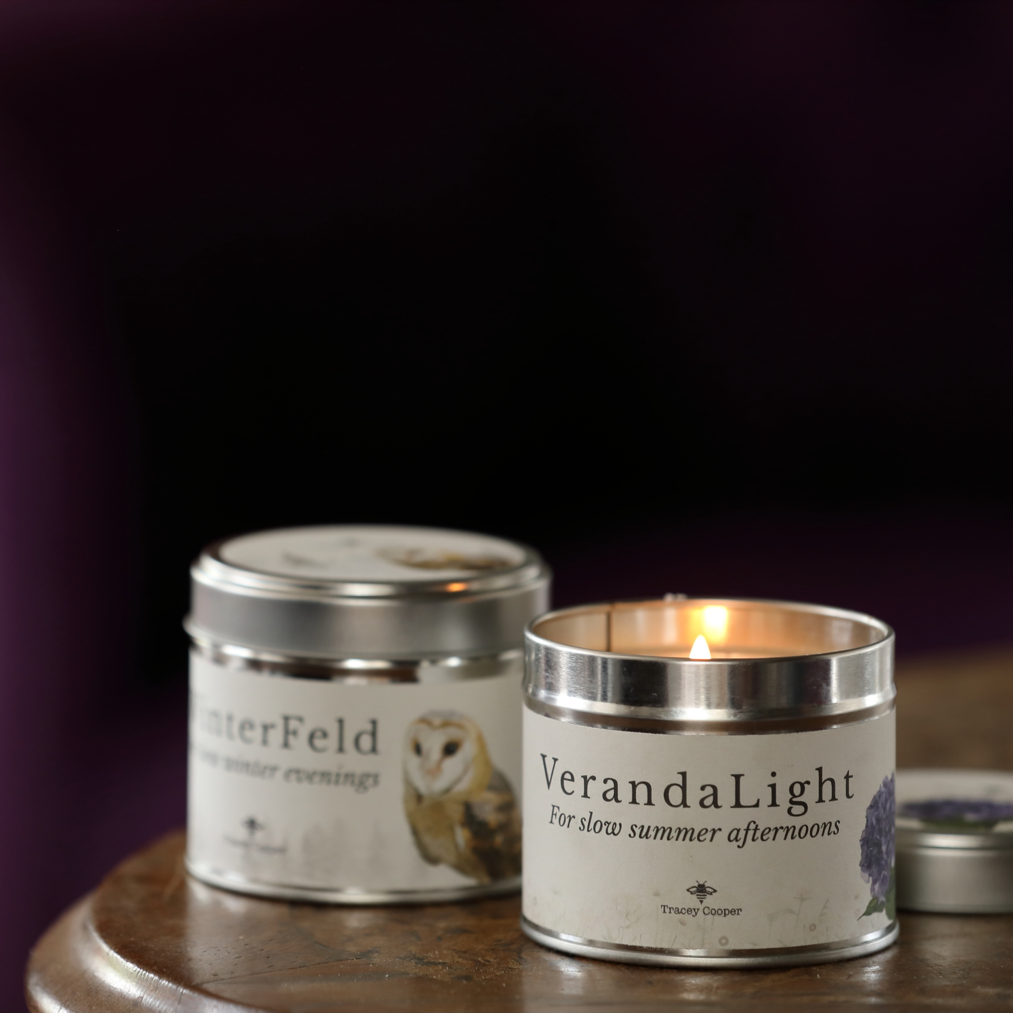 Two silver metal tin candles on a wooden surface against a dark purple background. One tin is closed with a lid, and the other is open with a lit flame, both featuring white labels with black text and botanical illustrations.