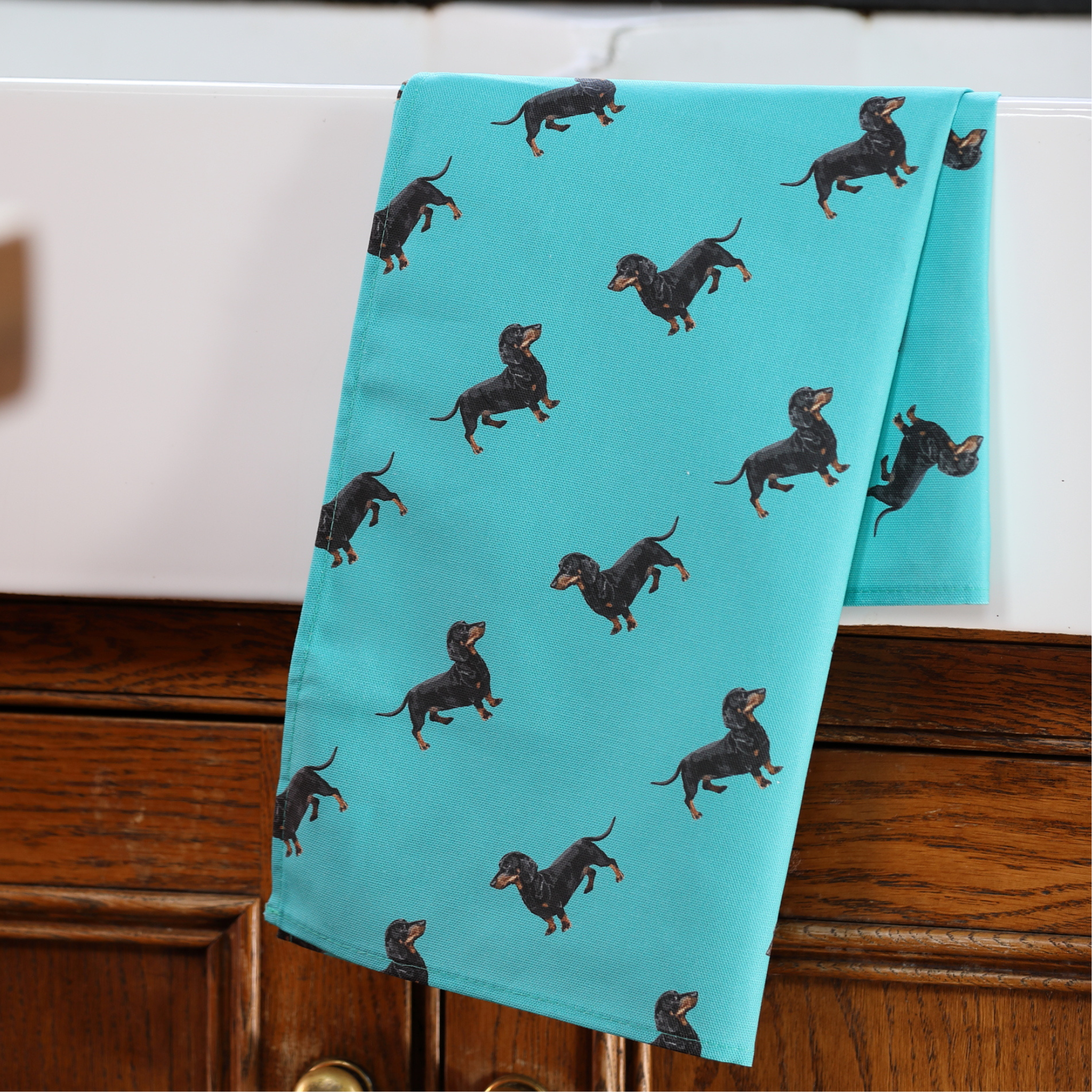 A turquoise kitchen towel with a repeating pattern of black and tan dachshunds, draped over a dark wood cabinet.