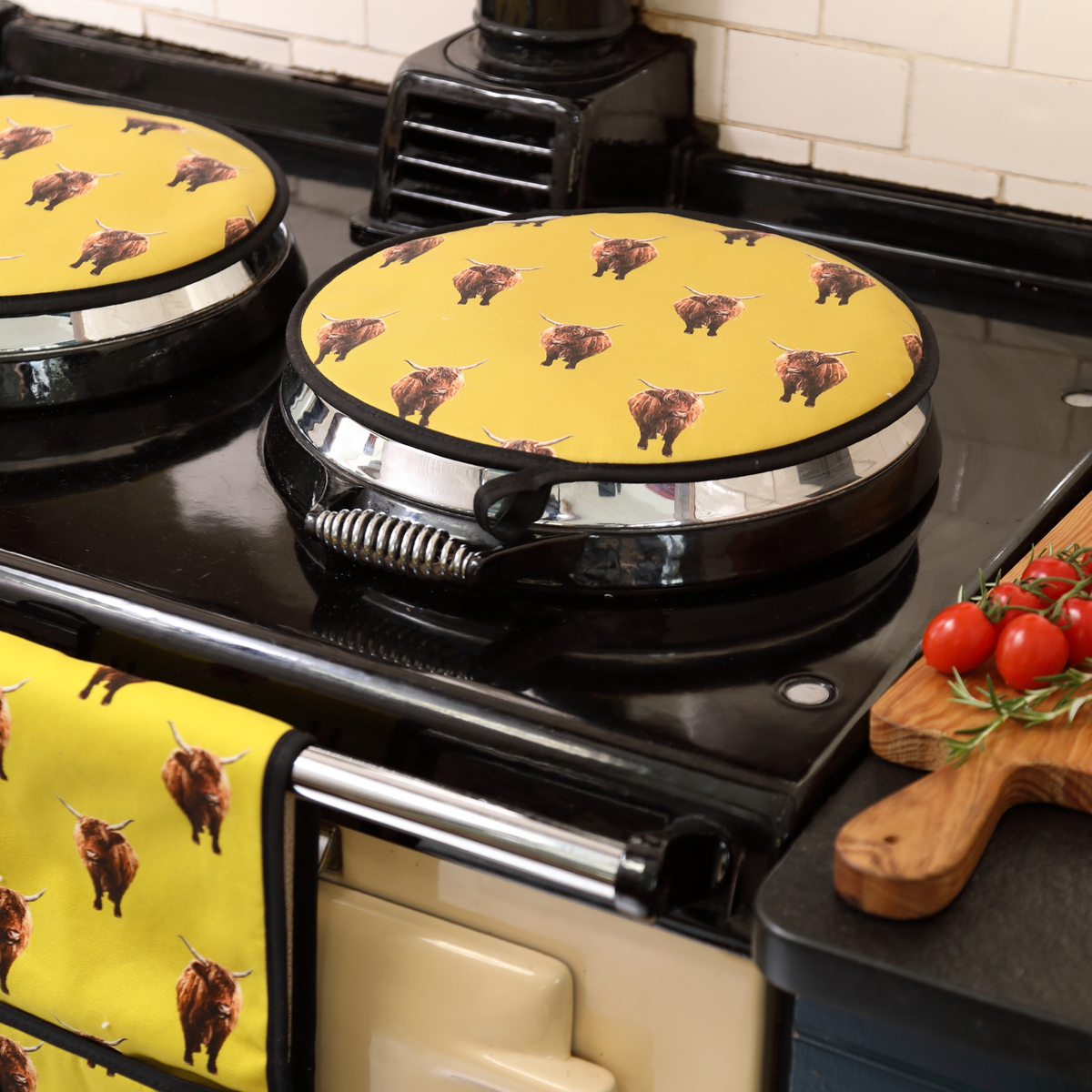 A yellow ovenglove and matching round AGA lid covers printed with a highland cow pattern, shown in a kitchen setting next to a black stove and a cutting board with tomatoes.