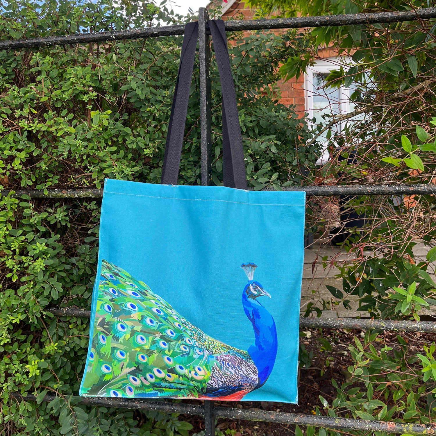 A tote bag with a illustration of a peacock on a teal background, hanging on a fence with bushes in the background.